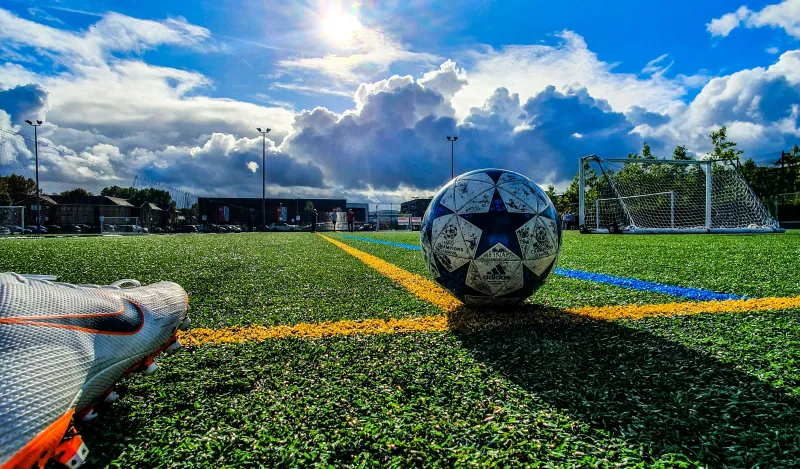 this is a stock image photo of a foot with a soccer cleat on, about to kick a soccer ball on a field on a bright sunny, blue sky with puffy white clouds day. 