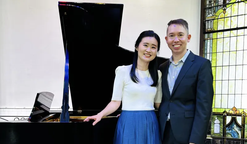 this is a smiling photo of NHCC music faculty, Grace Choi and Kevin Hobbs standing next to a piano. 