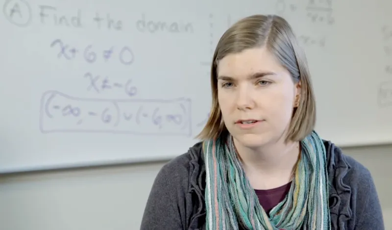 woman speaking with math problems on a white board behind her