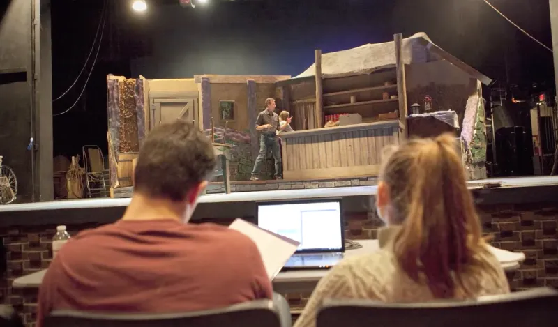 students next to a laptop with a stage and actors in front of them 