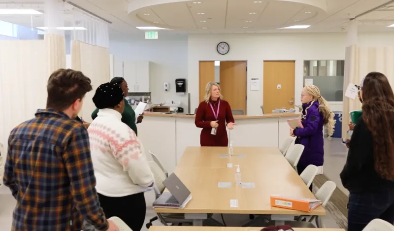 Female instructor speaking to a small group of students standing around a table in the nursing lab. 