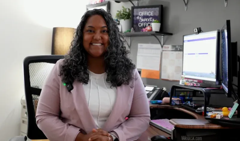 Woman sitting in her office smiling with a computer in the background. 