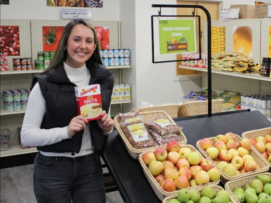 a photo of NHCC Social Worker, Ellie standing next to fruit in the Food Cupboard, smiling