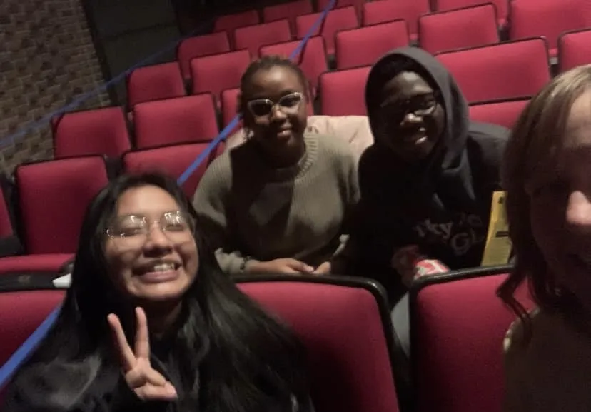a photo of three NHCC PTK students and their Advisor in the theater before seeing a show, with the lights dimmed. 