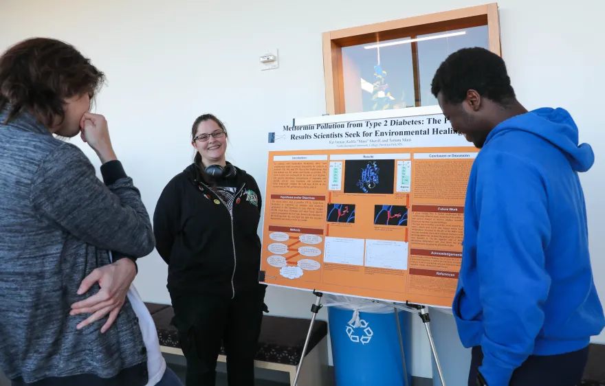 a photo of three students standing next to a poster of biology research. 