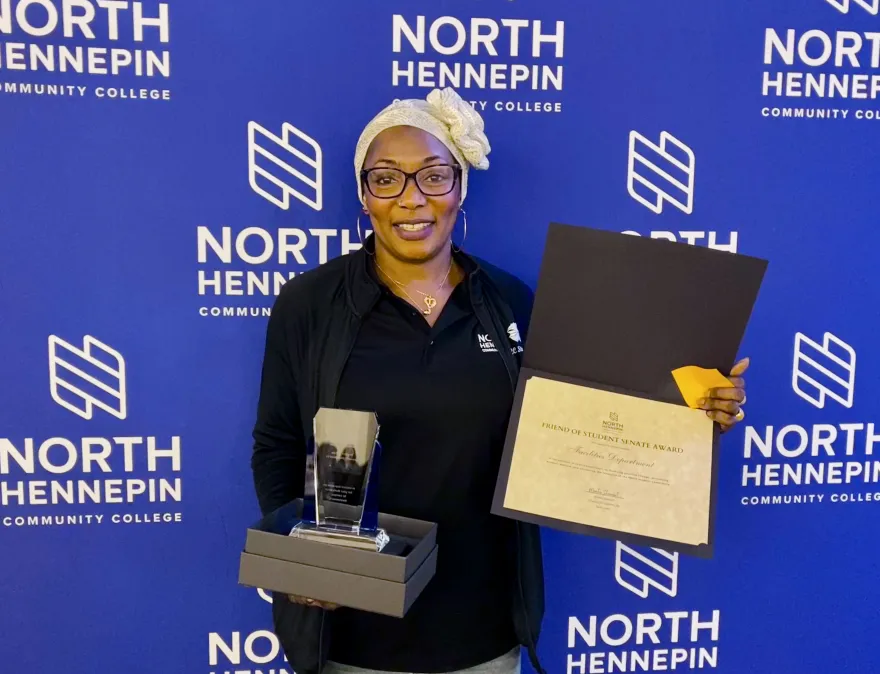This is a photo of NHCC staff member, Hawa standing in front of an NHCC logo "step and repeat" backdrop, she is smiling, holding a certificate and award, which is a plaque made of glass, after receiving recognition on behalf of her department at the Annual Student Life Awards Banquet. 