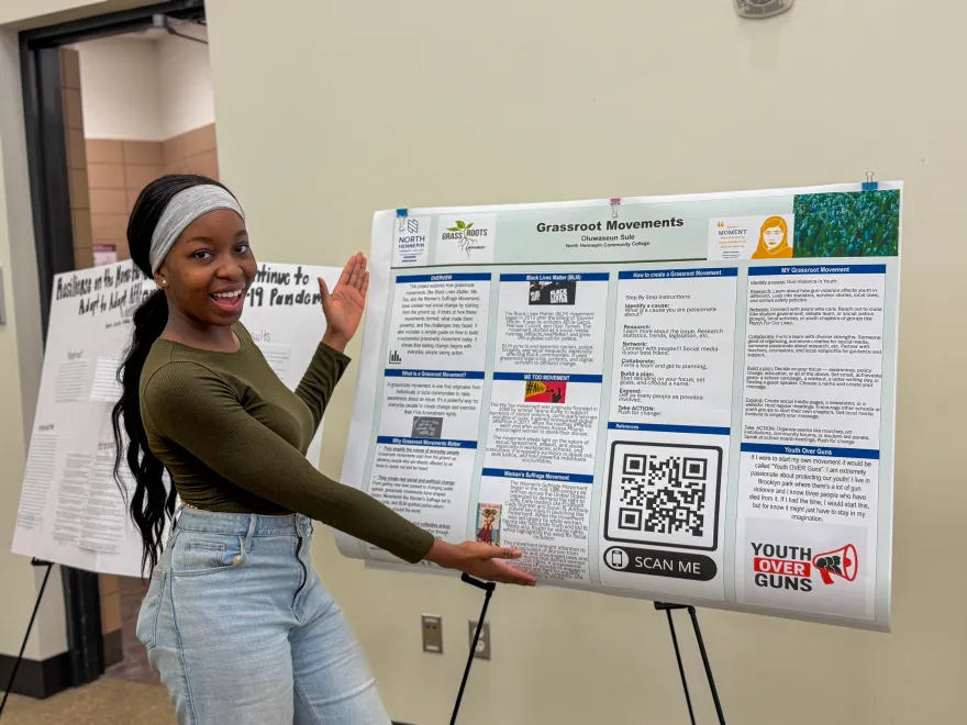 This is a photo of NHCC student and 2025 Commencement Speaker, Oluwaseun Sule standing with her arms out, gesturing to her research poster on display, smiling in Helling Hall on-campus for our annual NHCC+ research event. 