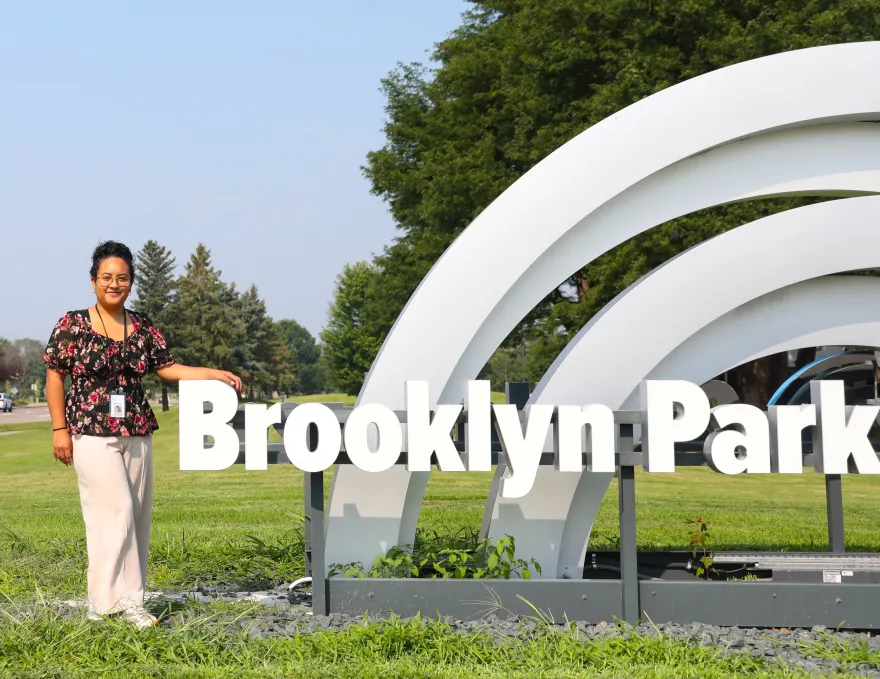 this is photo of NHCC graphic design alumni, Estefanny Zelaya standing in front of City Hall on the lawn, posing with the giant Brooklyn Park sign. 