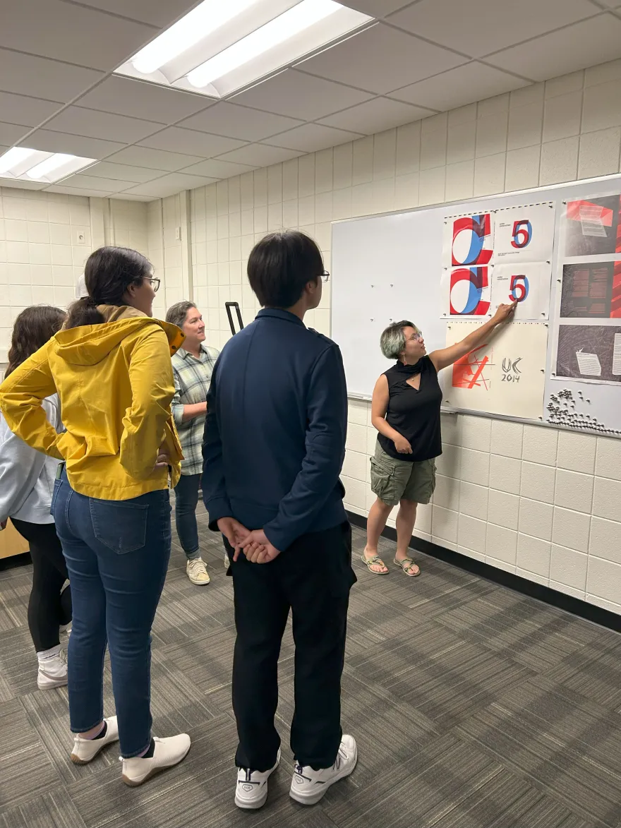 this is a photo of a student smiling standing next to a whiteboard in a Fine Arts Center classroom, pointing to a piece of art hanging on the wall. A few students and a teacher are gathered around the student in a semicircle, looking at the artwork during a graphic design critique practice. 