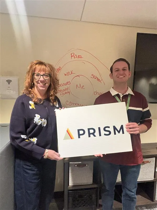 this is a photo of NHCC faculty, Amy Harms Hoad standing next to a male colleague smiling as they're both holding up a sign that reads, 'PRISM.'