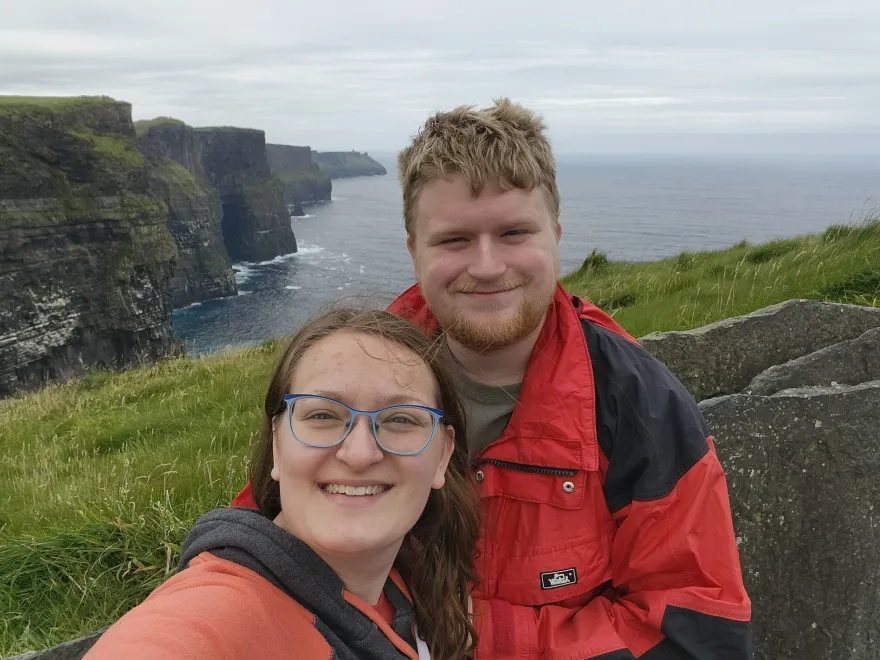 this is a photo of NHCC alumni, Maddi and Theo presumably on vacation near the coast of Ireland. There are cliffs, islands, greenery and ocean behind their smiling faces in the photo. 