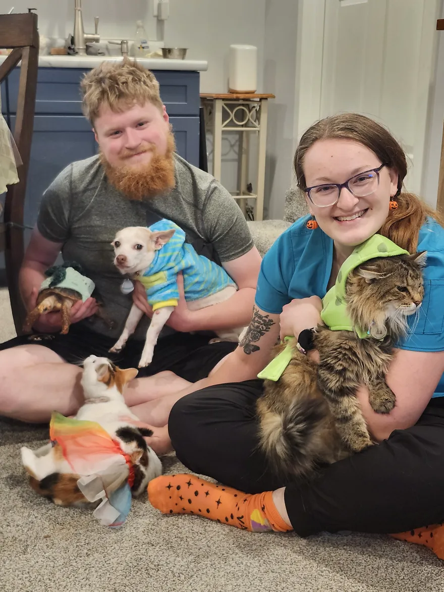 this is a photo of NHCC alumni, Theo and Maddi, sitting on the floor smiling for a photo with their pets. Pictured are two cats, a dog and a tortisse. 