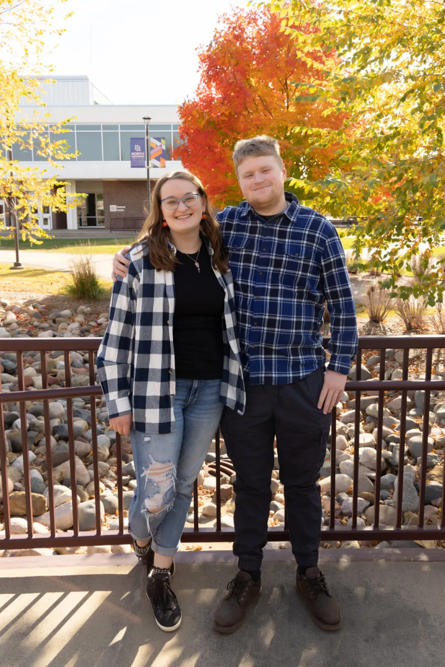 this is a photo of NHCC alumni Maddi and Theo posing on the bridge in our courtyard in the fall time with a red, yellow and orange leaved tree behind them. 