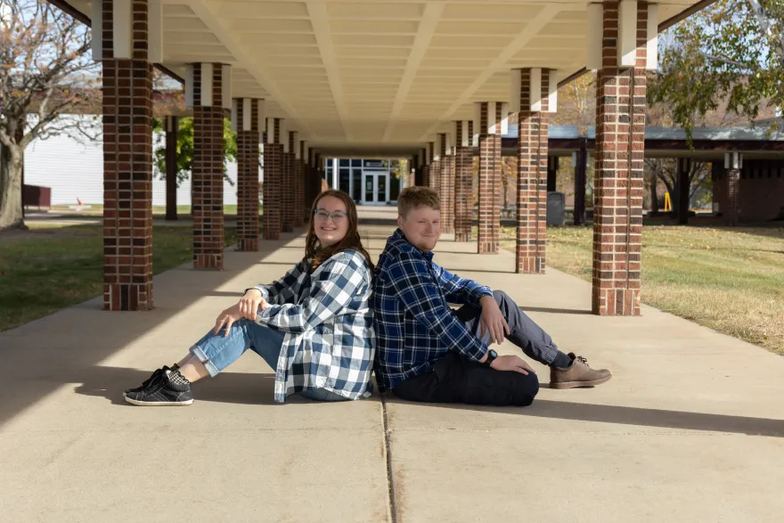 this is a photo of NHCC alumni, Maddi and Theo sitting back to back, on the concrete on NHCC's campus, under the brick pillars and breezeway, near the Campus Center building.