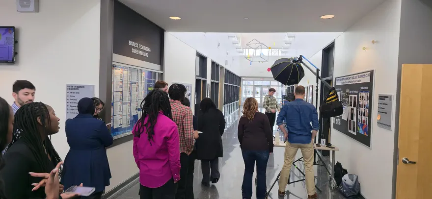 this is a photo from the CBT building hallway, taken while camera equipment was set up for a Kappa Beta Delta February Headshots event. This photo is a behind-the-scenes shot of students lined up about to get their photos taken, their backs are mostly to the camera. 