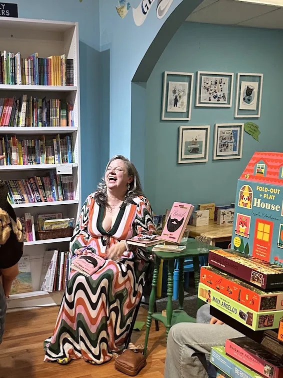 this is a photo of NHCC english faculty, Kelly Lundquist laughing, sitting in a bookstore called Roscoe Books next to a table with copies of her memoir on it, when she was on tour at a bookstore called Roscoe Books in Chicago, Illinois. 