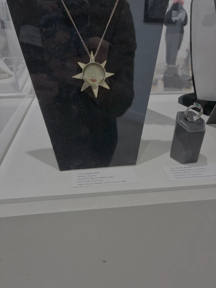 this is a photo of a gold necklace on a black velvet neck bust, in a clear plastic display case. The artist card reads, Title: Jeevan Jyoti Media: Jewelry Student's Name: Willow Johri Instructor: Erin Boe High School: Maple Grove Senior High. The gold necklace with a gold chain, features a sunshine with triangles, a face with eyes closed, red lips and a glass case in the center. 