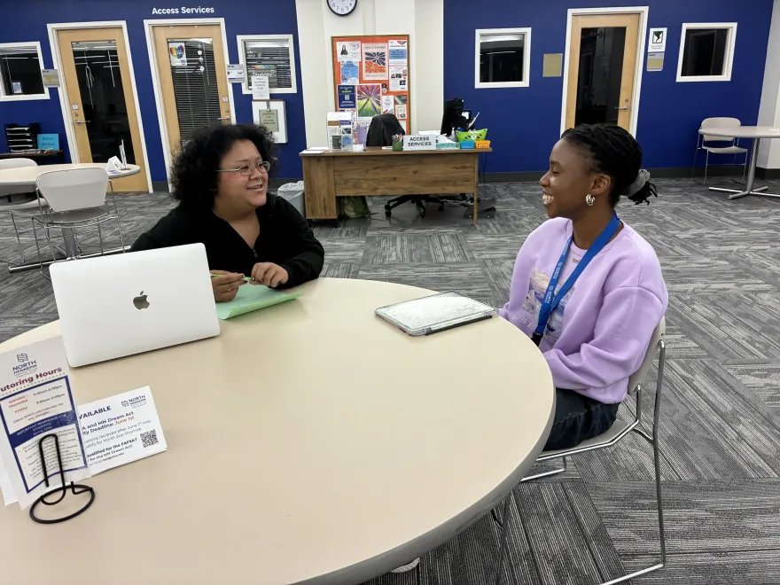 this is a photo of two female students, smiling, sitting at a table in the library. NHCC peer tutor, Ray Foss works with Biology student, Danista Kannah to edit a paper on the computer. 