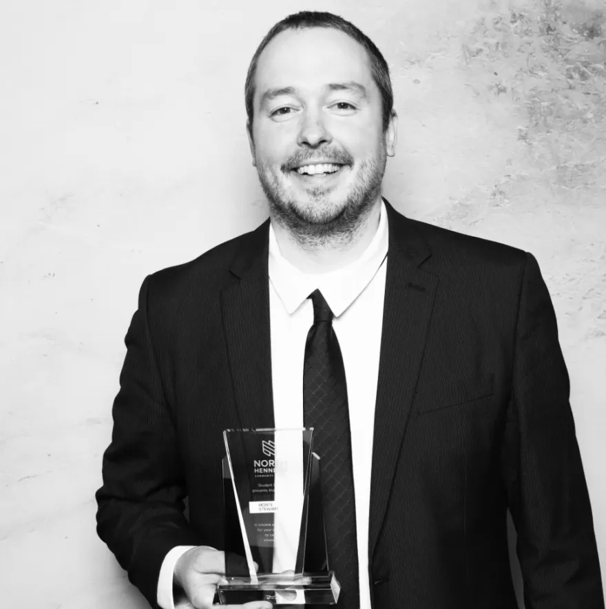 this is a black and white photobooth photo of NHCC staff, Monte Stewart smiling and holding up his Student Life Award certificate.
