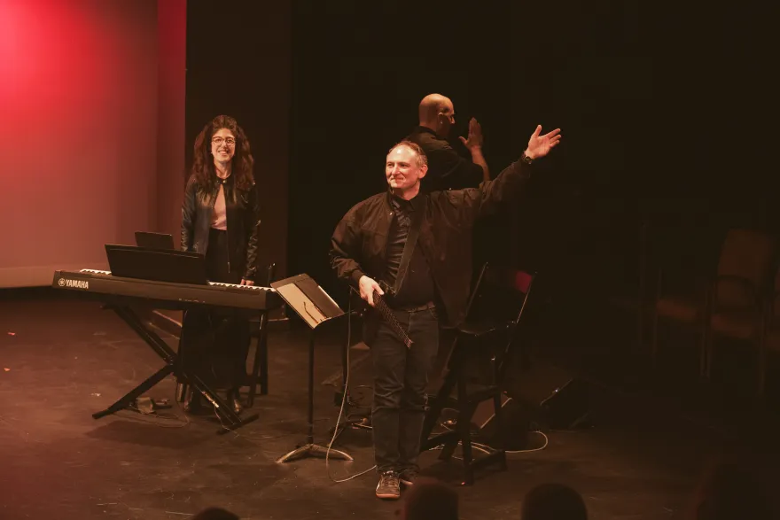 this is a photo of NHCC faculty, Gary Ruschman on stage, standing next to a piano player, with his arm out, almost as if bowing while being recognized by an enthusiastic audience.