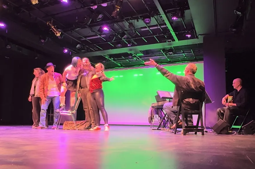 this is a photo of NHCC faculty, Gary Ruschman sitting in a chair on a stage facing five individuals, conducting the performance of Carmen.