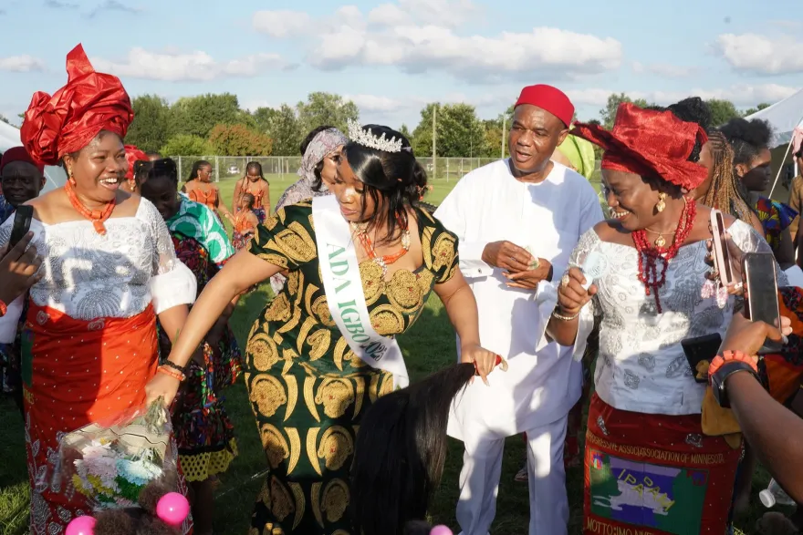 Attendees in traditional Igbo attire gather outdoors during IgboFest, smiling and dancing together as a woman wearing a sash participates in the celebration.
