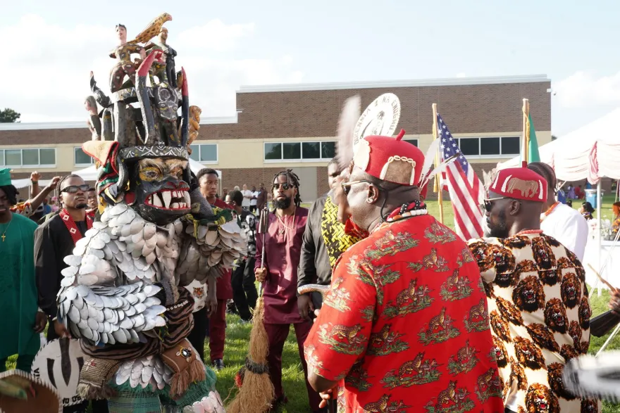 A performer wearing an elaborate traditional masquerade costume engages with attendees during an IgboFest cultural celebration, while spectators watch nearby.