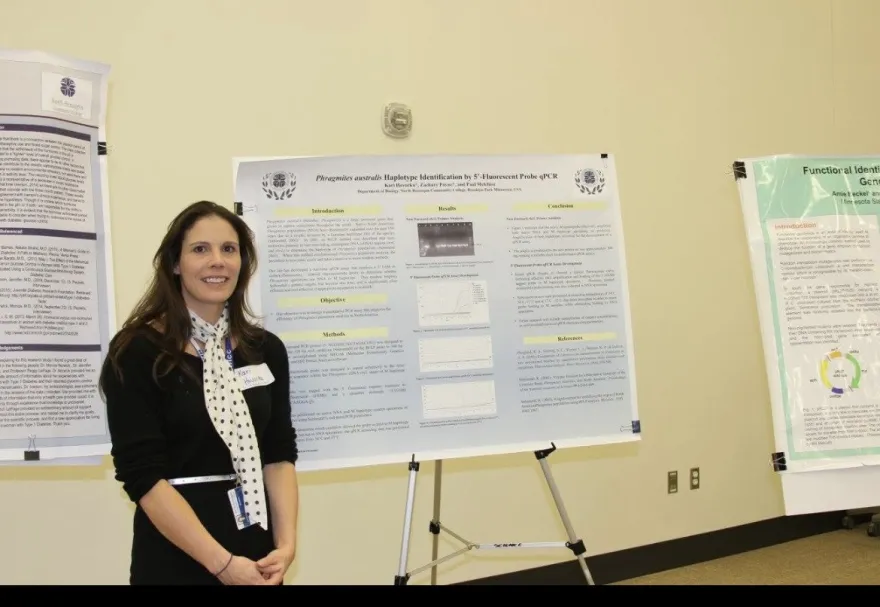 this is a smiling photo of NHCC graduate, Kari Struble, standing in the Grand Hall next to a poster with her scientific research on it. 