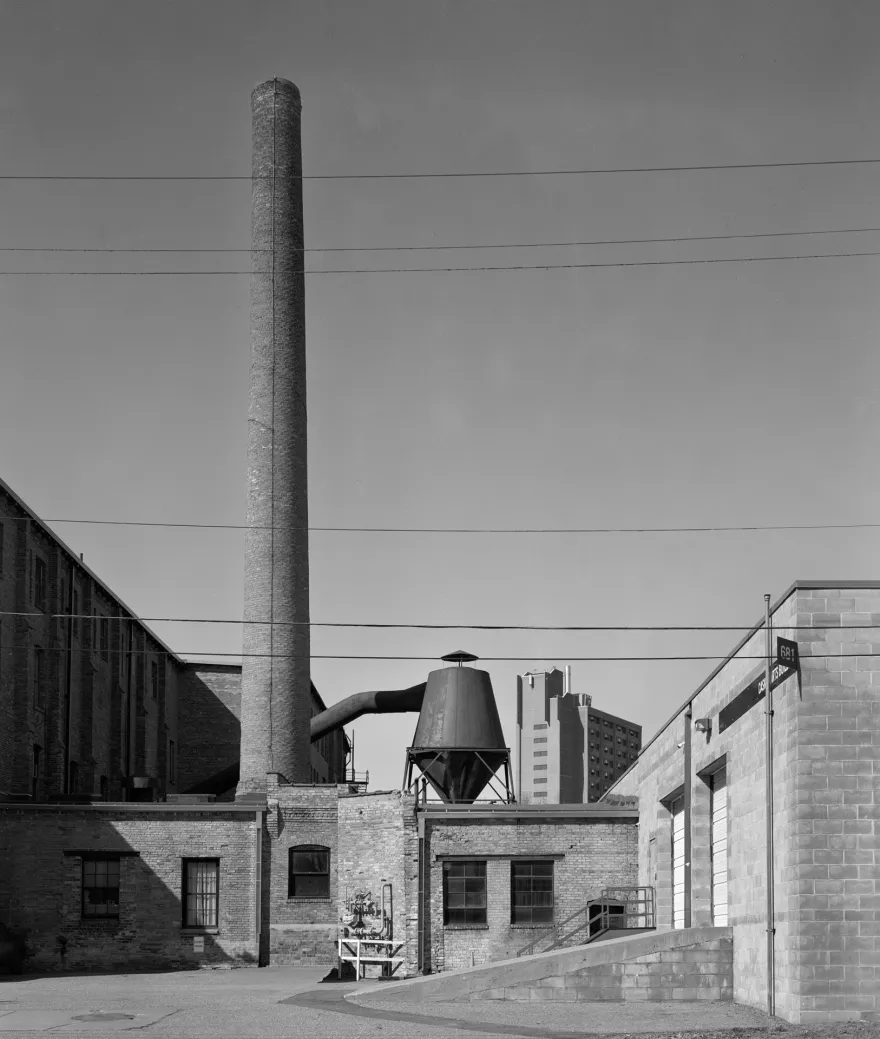 Black-and-white photograph of an industrial building complex, featuring a tall brick smokestack, metal hopper, exposed pipes, and several overhead utility wires against a clear sky.