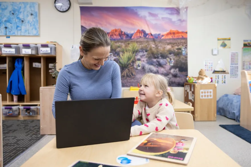 this is a photo of a mother sitting next to her daughter, they're both smiling and the mom is looking at her laptop and her daughter is seemingly tickling or touching her mom's leg. 
