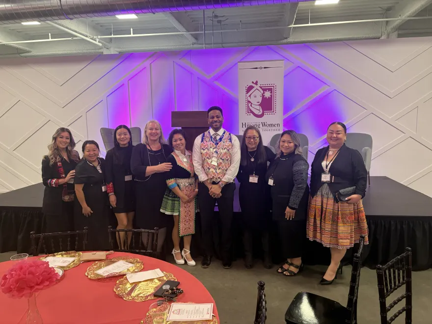 this is a smiling group photo of eight female NHCC staff members with Vice President of Student Affairs, Julio Vargas-Essex at a Hmong women Dinner Gala event. Julio is wearing a traditional Hmong money vest.