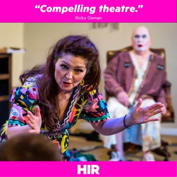 A woman in a bright dress facing the camera and talking to someone. A man sitting in a chair behind her.